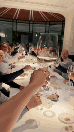 Un calice alzato per un brindisi sopra la tavola, con gli ospiti seduti lungo il tavolo sullo sfondo