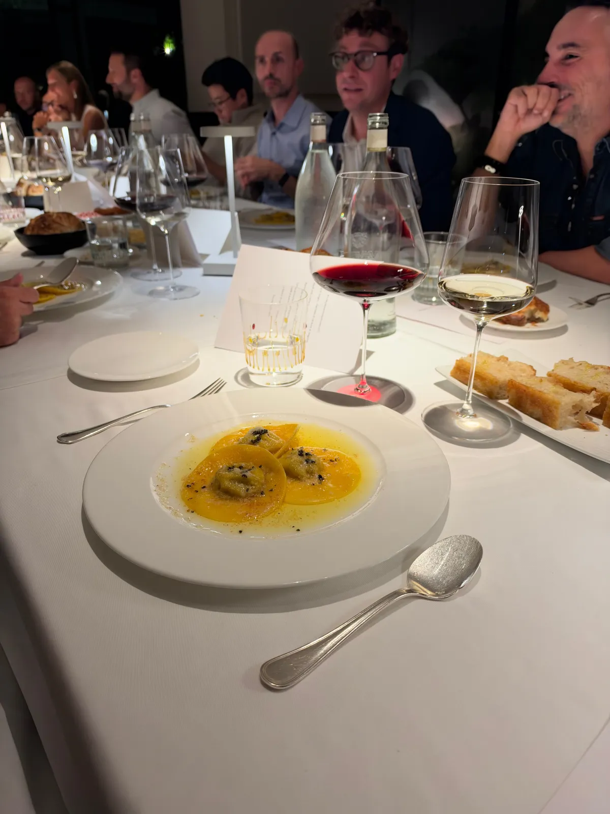 Un piatto di ravioli servito a tavola, con gli ospiti che chiacchierano e sorridono sullo sfondo